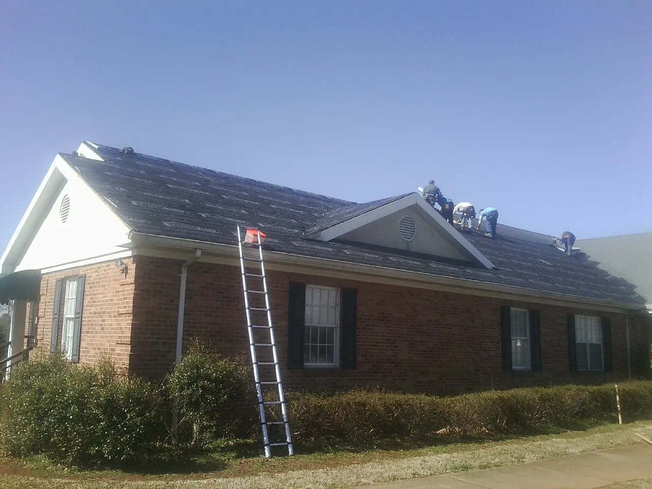 Crew tearing shingles off a brick ranch roof with a ladder set — Emergency Roof Repair work by our crew in Crescent Mills
