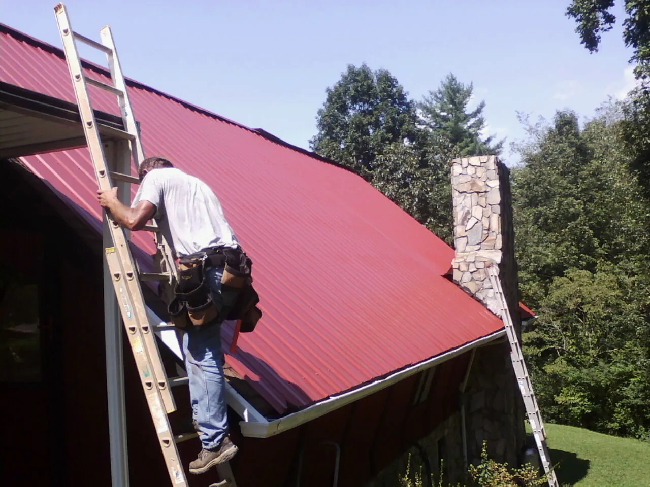 Roofer on a ladder beside a red metal roof and stone chimney — Soffit & Fascia Repair by Roofing Brothers in Crescent Mills